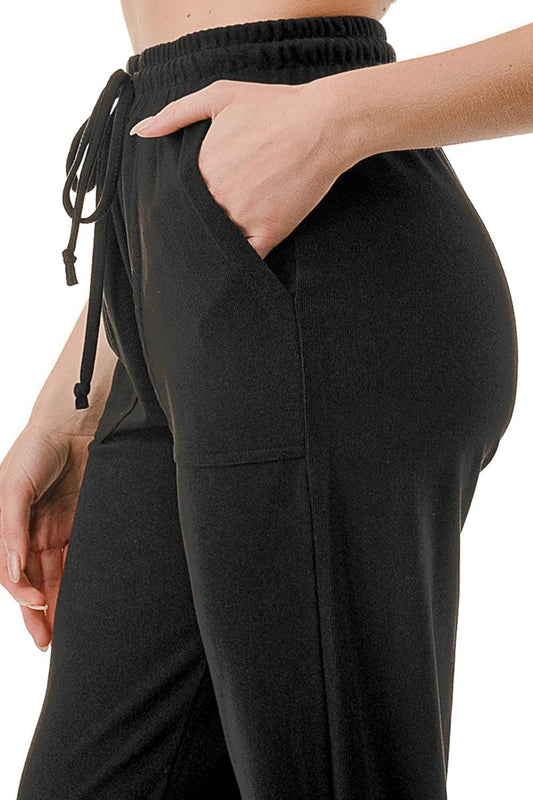 May include: Close-up of black sweatpants with a drawstring waistband and pockets. The pants are made of a soft, ribbed fabric. The drawstring is visible on the left side, and a hand is in the pocket. The pants appear casual and comfortable, suitable for loungewear or athletic activities.